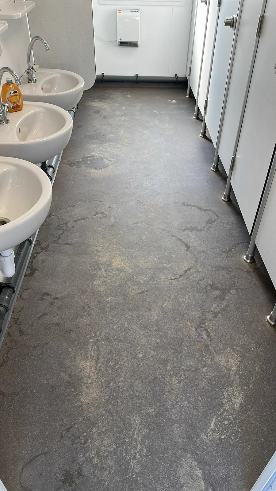 Sanitärraum mit sichtbaren Ablagerungen auf dem grauen Boden, Waschbecken links und Kabinen rechts.